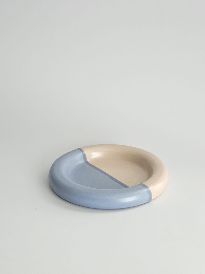 May include: A round ceramic dish featuring a smooth, glossy finish. The surface is divided into two distinct sections, with one half colored in a soft, muted blue and the other half in a creamy, off-white tone, creating a simple, balanced design.