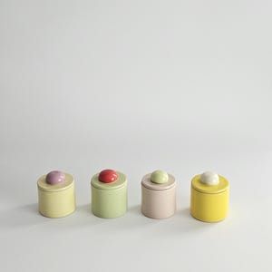 May include: Four cylindrical ceramic canisters with lids, each topped with a small rounded knob. The canisters are colored pale yellow, light green, soft pink, and bright yellow, featuring knobs in lavender, red, pale green, and white.