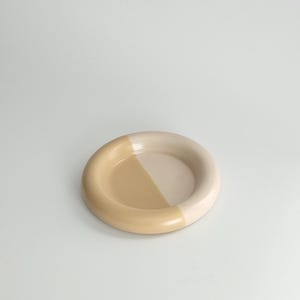 May include: A circular ceramic dish featuring a smooth, glossy finish. The design is divided into two distinct color sections, with one half in a soft cream tone and the other half in a warm, muted tan shade, creating a balanced two-tone appearance.
