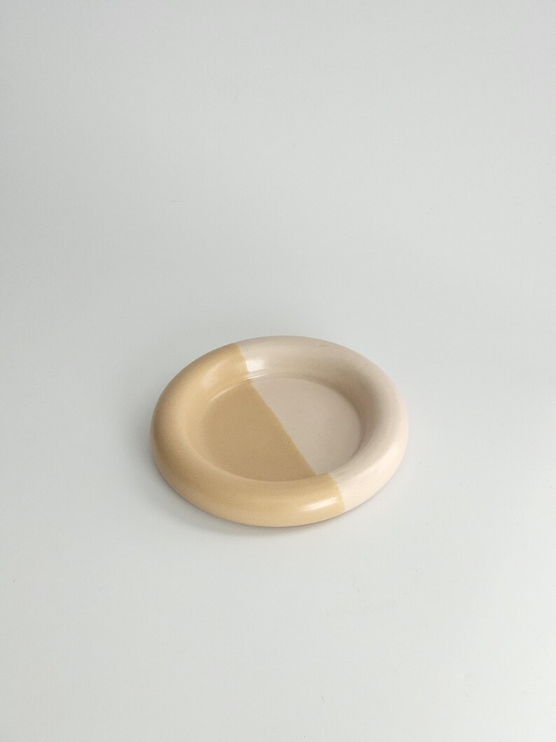May include: A circular ceramic dish featuring a smooth, glossy finish. The design is divided into two distinct color sections, with one half in a soft cream tone and the other half in a warm, muted tan shade, creating a balanced two-tone appearance.