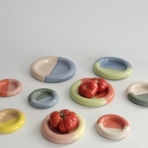 May include: A collection of small, round ceramic plates in various pastel and muted color combinations, including blue, pink, yellow, green, and peach. Two of the plates hold fresh, red tomatoes. The plates feature a smooth, glossy finish.