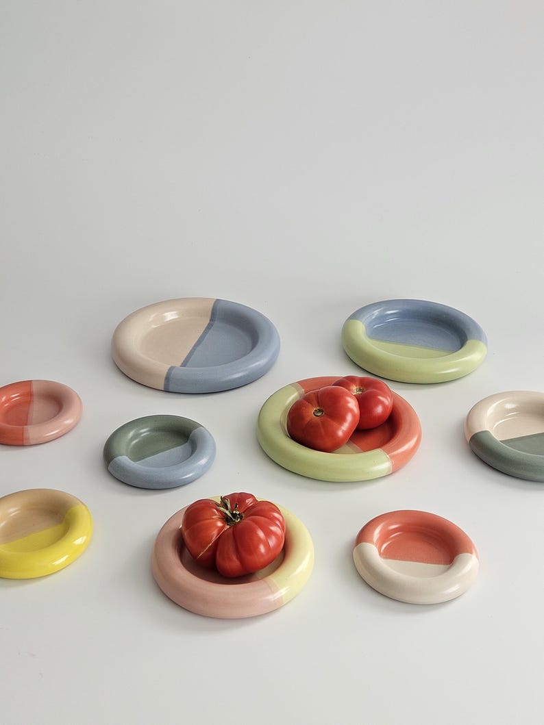 May include: A collection of small, round ceramic plates in various pastel and muted color combinations, including blue, pink, yellow, green, and peach. Two of the plates hold fresh, red tomatoes. The plates feature a smooth, glossy finish.