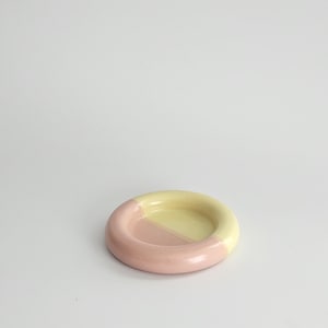 May include: A small, circular ceramic dish featuring a smooth, glazed finish. The object is divided into two distinct color sections, with one half colored in a soft pastel pink and the other half in a pale, creamy yellow tone.