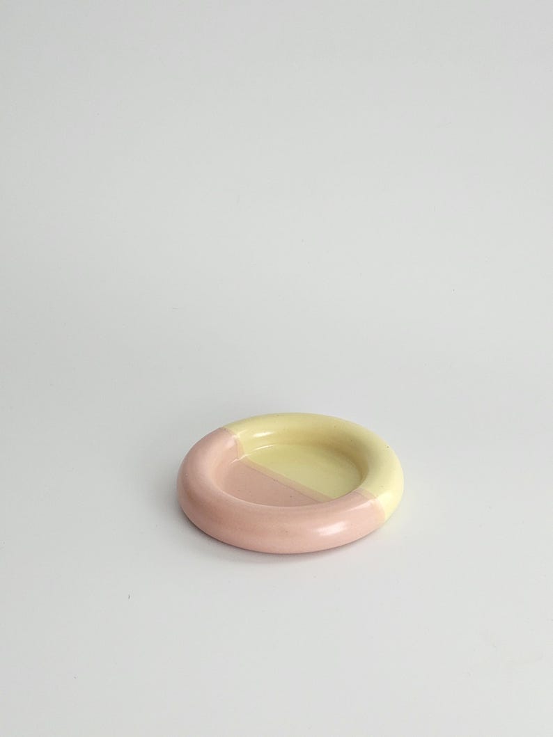May include: A small, circular ceramic dish featuring a smooth, glazed finish. The object is divided into two distinct color sections, with one half colored in a soft pastel pink and the other half in a pale, creamy yellow tone.