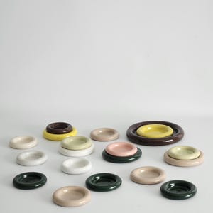 May include: A collection of small, circular, concave ceramic dishes in various colors including white, cream, pale yellow, soft pink, dark green, and deep brown. The dishes are arranged in a scattered pattern, with some stacked on top of each other.