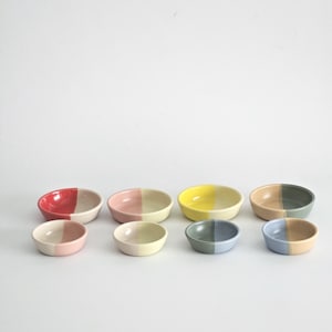 May include: A collection of eight small, ceramic bowls. Each bowl is divided into two color sections, featuring combinations of red, pink, yellow, gray, and blue. The bowls are arranged on a white surface.