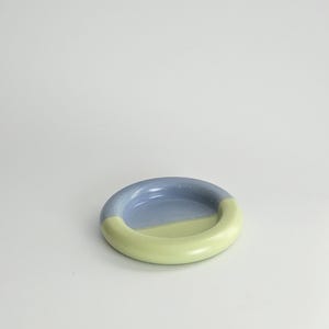 May include: A small, circular ceramic dish with a smooth, glossy finish. The dish features a two-tone color scheme, with one half colored in a soft periwinkle blue and the other half in a pale, muted lime green, separated by a clean line.