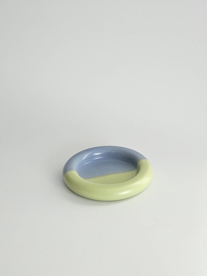 May include: A small, circular ceramic dish with a smooth, glossy finish. The dish features a two-tone color scheme, with one half colored in a soft periwinkle blue and the other half in a pale, muted lime green, separated by a clean line.