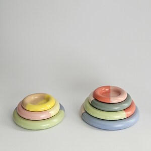 May include: Two stacks of circular ceramic objects. The left stack features three rings in yellow, pink, and light green. The right stack consists of four rings in coral, sage green, light green, and periwinkle blue, each with a smooth finish.