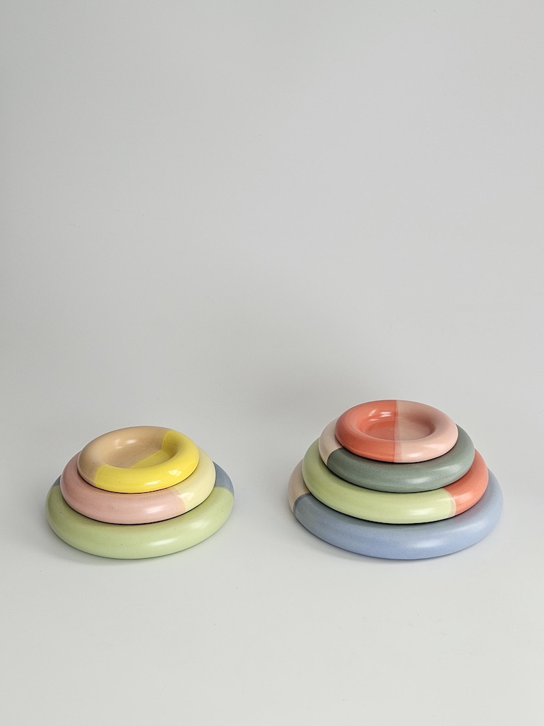 May include: Two stacks of circular ceramic objects. The left stack features three rings in yellow, pink, and light green. The right stack consists of four rings in coral, sage green, light green, and periwinkle blue, each with a smooth finish.