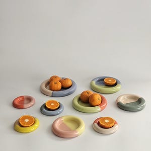 May include: A collection of eight small, round, ceramic plates in various pastel color combinations, including pink, blue, green, and yellow. Several plates hold whole or halved oranges, showcasing the smooth, glossy finish of the dishware.