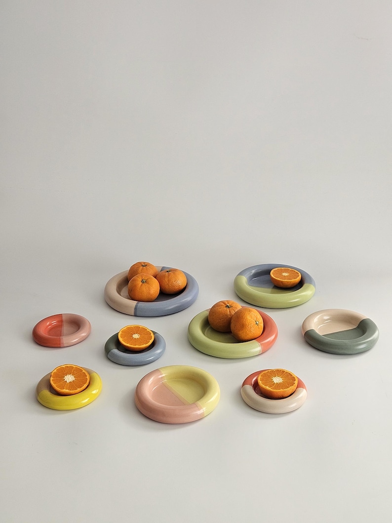 May include: A collection of eight small, round, ceramic plates in various pastel color combinations, including pink, blue, green, and yellow. Several plates hold whole or halved oranges, showcasing the smooth, glossy finish of the dishware.