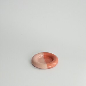 May include: A small, circular ceramic dish with a smooth, glossy finish. The dish features a distinct two-tone color scheme, with one half in a soft, pale pastel pink and the other half in a deeper, saturated terracotta or coral orange shade.