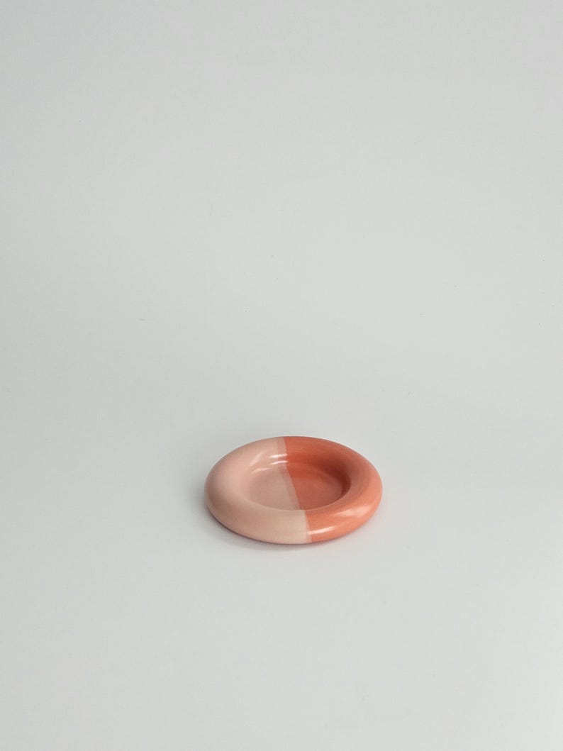 May include: A small, circular ceramic dish with a smooth, glossy finish. The dish features a distinct two-tone color scheme, with one half in a soft, pale pastel pink and the other half in a deeper, saturated terracotta or coral orange shade.