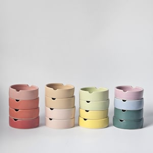 May include: A set of six ceramic ashtrays stacked in different colors, including red, orange, yellow, green, blue, and pink. The ashtrays are all the same size and shape, and they are arranged in a row on a white background.