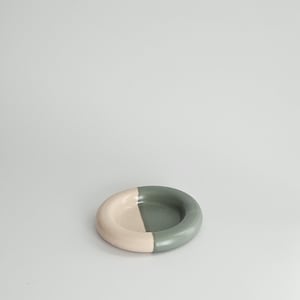 May include: A small, circular ceramic dish featuring a smooth, matte finish. The bowl is divided into two distinct color sections, with one half colored in a soft, pale beige and the other half in a muted, earthy sage green tone.
