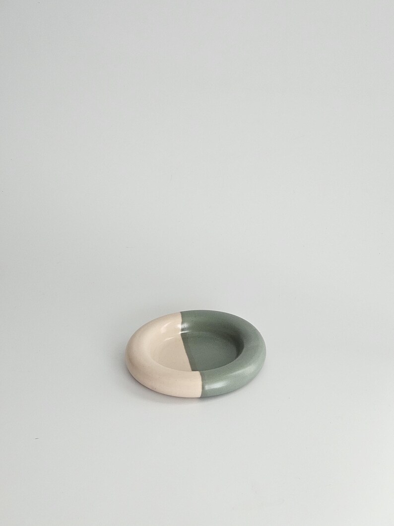 May include: A small, circular ceramic dish featuring a smooth, matte finish. The bowl is divided into two distinct color sections, with one half colored in a soft, pale beige and the other half in a muted, earthy sage green tone.