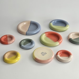 May include: A collection of nine round ceramic dishes in various sizes, each featuring a two-tone color scheme. One dish is cream-colored with the text BAD DOGS! printed in black. The other dishes display combinations of blue, pink, yellow, green, and orange.