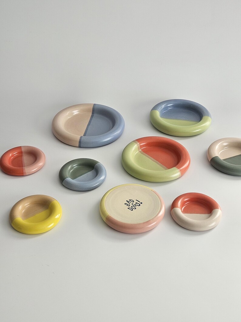 May include: A collection of nine round ceramic dishes in various sizes, each featuring a two-tone color scheme. One dish is cream-colored with the text BAD DOGS! printed in black. The other dishes display combinations of blue, pink, yellow, green, and orange.