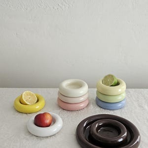 May include: A collection of smooth, circular ceramic bowls with thick, rounded rims in various colors including yellow, white, cream, pink, light green, blue, and dark brown. Some bowls contain pieces of fruit like a lemon, lime, and a nectarine.