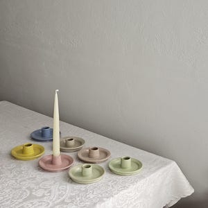 May include: A collection of seven small, round ceramic candle holders in various pastel colors including yellow, pink, blue, beige, and green. One pink candle holder contains a single, tall, slender, off-white taper candle standing upright.