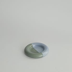 May include: A small, circular ceramic dish with a smooth, glazed finish. The object features a two-tone color scheme, with one half in a muted sage green and the other half in a soft, pale periwinkle blue, meeting in a clean, diagonal line.