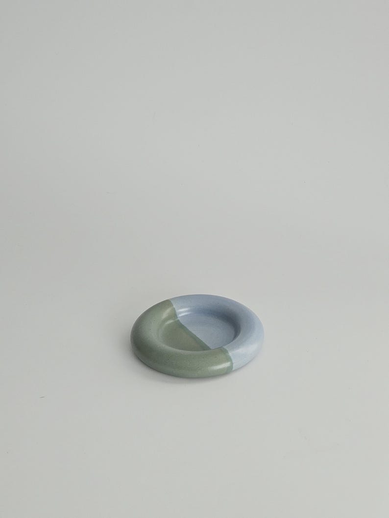 May include: A small, circular ceramic dish with a smooth, glazed finish. The object features a two-tone color scheme, with one half in a muted sage green and the other half in a soft, pale periwinkle blue, meeting in a clean, diagonal line.