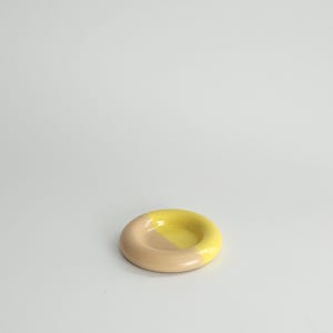 May include: A small, circular ceramic dish with a smooth, glossy finish. The object features a two-tone color scheme, with one half colored in a soft, muted beige and the other half in a bright, vibrant yellow, creating a distinct contrast.