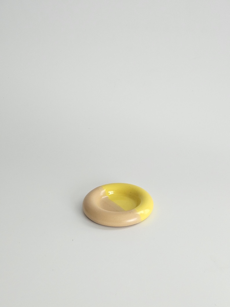 May include: A small, circular ceramic dish with a smooth, glossy finish. The object features a two-tone color scheme, with one half colored in a soft, muted beige and the other half in a bright, vibrant yellow, creating a distinct contrast.