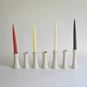 May include: A collection of six off-white ceramic candle holders, each holding a different colored taper candle: red, yellow, and green. The holders have a flared base and a narrow neck, creating a minimalist aesthetic.