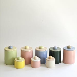 May include: A collection of ceramic jars in various sizes and pastel colors, including green, pink, yellow, blue, and cream. Each jar has a matching lid with a rounded knob. The jars are arranged in a row against a white background.