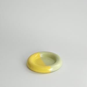 May include: A small, circular ceramic dish with a smooth, glossy finish. The dish features a two-tone color scheme, with one half colored in a bright, vibrant yellow and the other half in a soft, muted sage green, creating a distinct contrast.