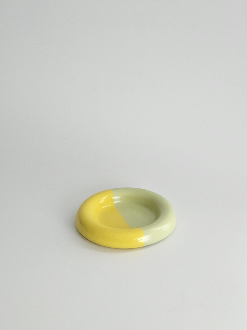 May include: A small, circular ceramic dish with a smooth, glossy finish. The dish features a two-tone color scheme, with one half colored in a bright, vibrant yellow and the other half in a soft, muted sage green, creating a distinct contrast.