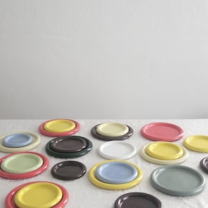 May include: A collection of round ceramic plates and saucers in various solid colors including yellow, pink, light blue, sage green, white, dark gray, and dark green. The pieces feature a raised circular center and a smooth, glossy finish.