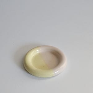 May include: A small, round ceramic dish with a smooth, glossy finish. The surface is divided into two distinct color sections, featuring a soft pastel yellow on one side and a muted, pale pink on the other, with a raised, rounded rim.