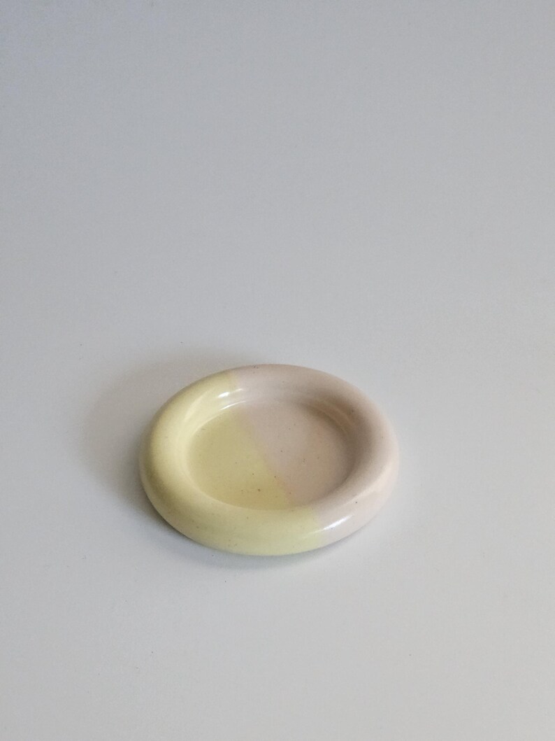 May include: A small, round ceramic dish with a smooth, glossy finish. The surface is divided into two distinct color sections, featuring a soft pastel yellow on one side and a muted, pale pink on the other, with a raised, rounded rim.