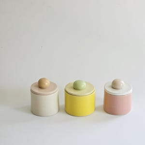 May include: Three small, cylindrical ceramic jars with lids. The jars are painted in cream, yellow, and pink, each with a matching lid and a spherical knob. The jars are arranged in a row against a white background.