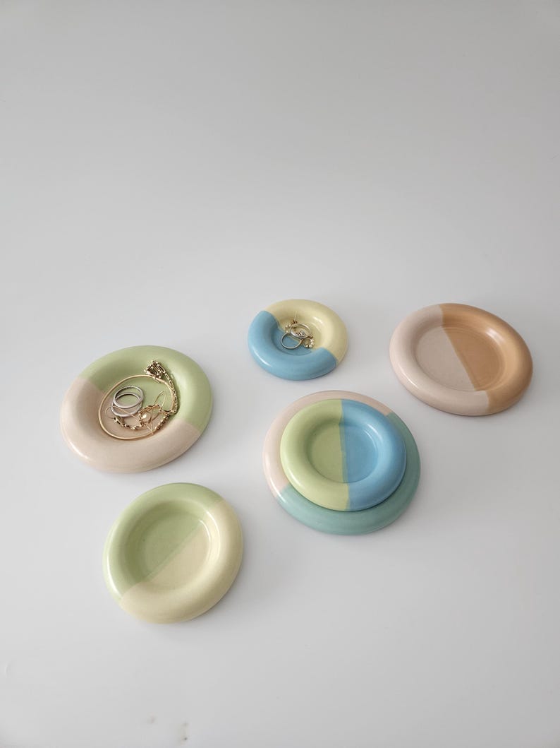 May include: A collection of five circular ceramic trinket dishes in various pastel color combinations including sage green, light blue, and soft peach. One dish holds a gold chain and rings, while another contains a small gold earring.