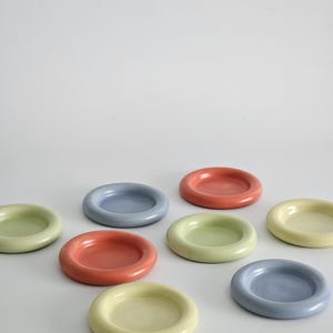 May include: A collection of seven small, circular ceramic dishes arranged in a scattered pattern. The dishes feature a smooth, glossy finish and come in a variety of pastel colors, including soft blue, pale yellow, and muted coral-orange tones.