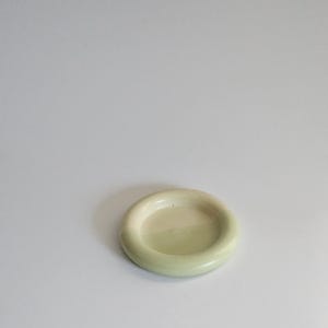 May include: A small, circular ceramic dish with a smooth, glossy finish in a pale, muted sage green color. The dish features a thick, rounded rim that encircles a shallow, flat center, creating a simple, uniform, and functional design.