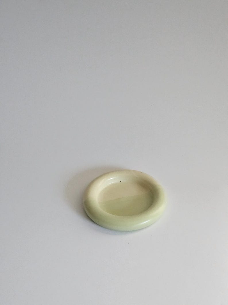 May include: A small, circular ceramic dish with a smooth, glossy finish in a pale, muted sage green color. The dish features a thick, rounded rim that encircles a shallow, flat center, creating a simple, uniform, and functional design.