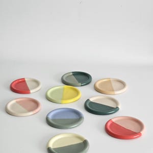 May include: A collection of eight circular ceramic coasters, each featuring a two-toned color scheme. The coasters display various combinations of red, pink, yellow, light green, dark green, blue, and beige, arranged in a semi-circle pattern.
