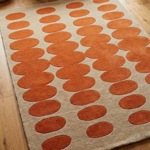 Könnte beinhalten: Ein rechteckiger Teppich mit einem sich wiederholenden Muster aus orangefarbenen ovalen Formen auf beigem Hintergrund. Der Teppich besteht aus einem weichen, strukturierten Material und liegt auf einem Holzboden. Das Design des Teppichs verleiht dem Raum eine moderne Note.