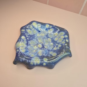 May include: A ceramic dish with an irregular shape. The dish has a glossy finish with a mix of blue, yellow, and white hues. The edge has a wavy design, enhancing its artistic look. This decorative item is suitable for serving or display, measuring approximately 20cm across.