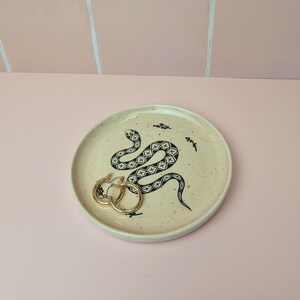 May include: A round, speckled ceramic dish with a black snake illustration and gold hoop earrings. The dish is beige with black speckles and a black snake design. The earrings are gold and are resting on the dish.