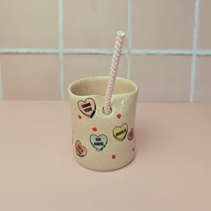 May include: A speckled, pale pink ceramic cup with a pink and white striped straw. The cup is decorated with colourful heart-shaped candies with phrases like "Kiss You" and "Be Mine".