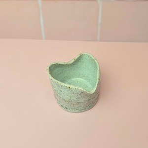 May include: A handmade ceramic heart-shaped dish with a speckled, pale green glaze. The dish has an organic shape and a slightly rough texture. The interior is a lighter shade of green. It is likely used for serving or as a decorative item.