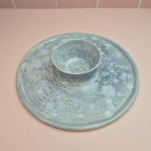 May include: A circular, pale blue ceramic serving platter with an integrated central bowl. The platter and bowl feature a speckled, textured finish with touches of green and white. Ideal for serving snacks or dips, the dish is a stylish addition.