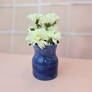 May include: A small, blue ceramic vase with a textured finish holds a bouquet of white chrysanthemums. The vase has a rounded shape and a narrow neck. The flowers are in full bloom, with many petals. The background is a pink surface.