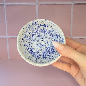 May include: A small, round ceramic bowl with a white base and a speckled pattern of dark blue paint splatters across the interior surface. The bowl features a smooth, glossy finish and a simple, circular rim, showcasing a handcrafted aesthetic.